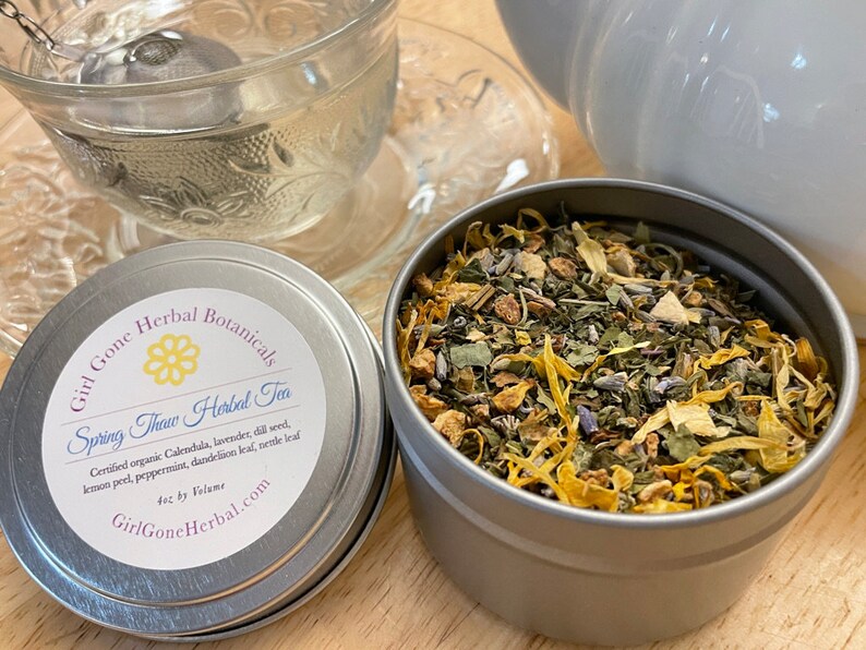 May include: A silver tin with a white label that reads "Girl Gone Herbal Botanicals Spring Thaw Herbal Tea" and lists the ingredients. The tin is filled with a blend of dried herbs, including lavender, calendula, lemon peel, and peppermint.