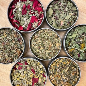 May include: Seven metal tins filled with different types of dried herbs and flowers. The tins are arranged in a circle on a wooden surface. The herbs and flowers are a mix of green, brown, yellow, and red.