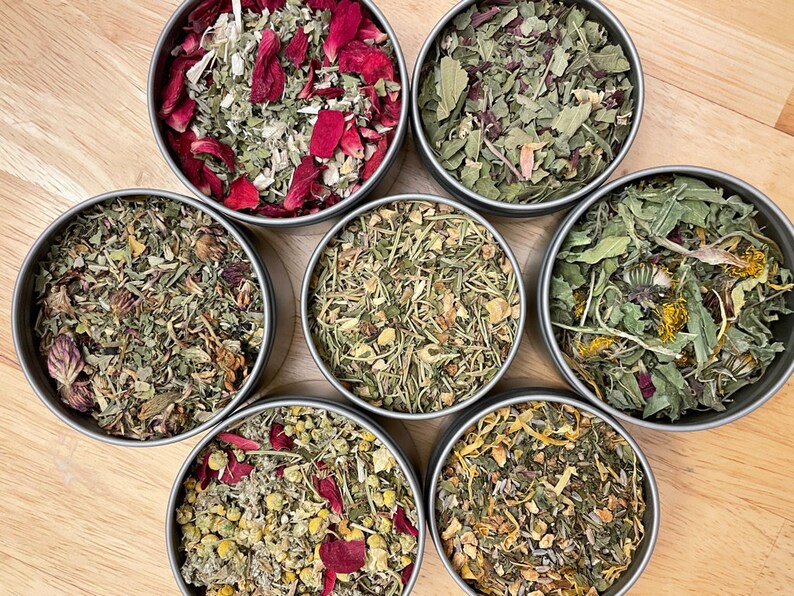 May include: Seven metal tins filled with different types of dried herbs and flowers. The tins are arranged in a circle on a wooden surface. The herbs and flowers are a mix of green, brown, yellow, and red.