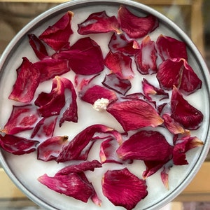 May include: A round, silver-colored container filled with a white substance and adorned with deep red rose petals. The petals are scattered across the surface, creating a visually appealing arrangement. This item is likely a scented candle.