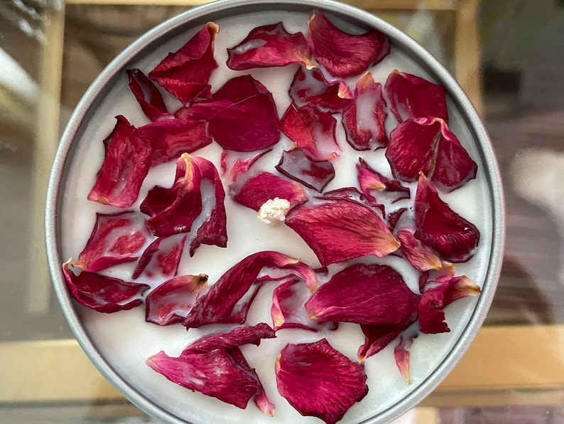 May include: A round, silver-colored container filled with a white substance and adorned with deep red rose petals. The petals are scattered across the surface, creating a visually appealing arrangement. This item is likely a scented candle.