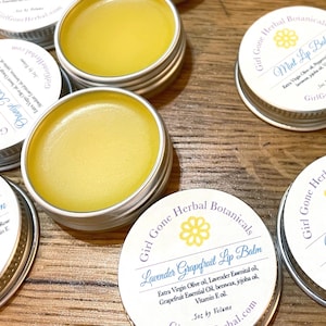 May include: Several small, round metal tins filled with yellow lip balm are arranged on a wooden surface. The tins have white lids with text and a floral design. The labels read "Girl Gone Herbal Botanicals" and list ingredients like olive oil and vitamin E.