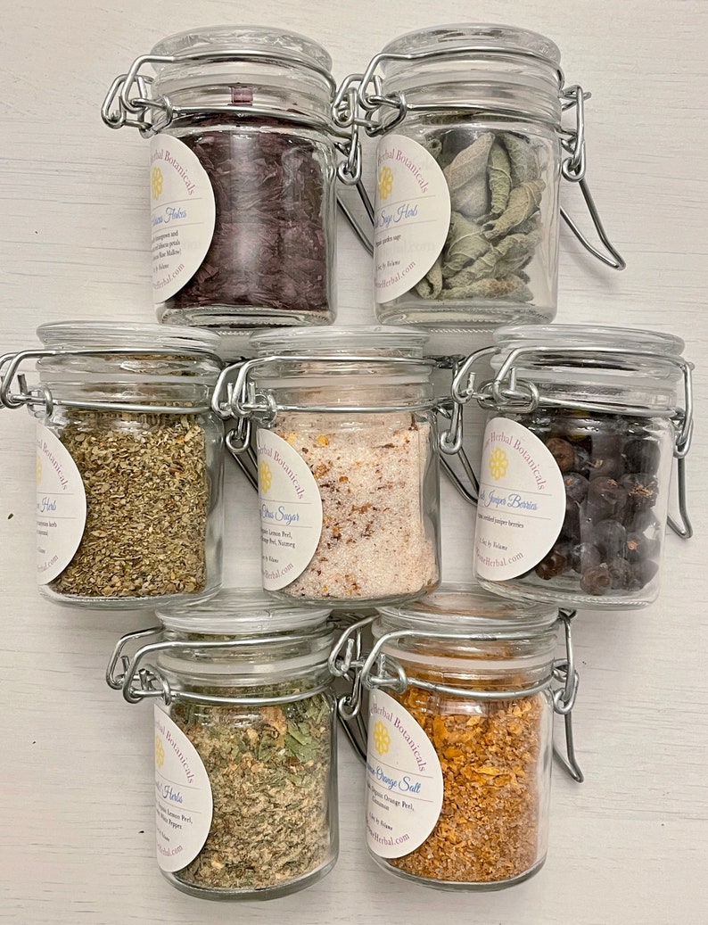 May include: Seven glass jars with metal lids containing various dried herbs and spices. The jars are labelled with the text "Herbal Botanicals" and the name of the herb or spice. The herbs and spices include: Hibiscus Flowers, Sage Herb, Oregano Herb, Citrus Sugar, Juniper Berries, Lemon Orange Salt.