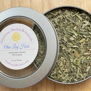 Può includere: Una scatola di latta argentata con un coperchio trasparente contenente foglie di olivo essiccate. L'etichetta sulla scatola recita "Girl Gone Herbal Botanicals Olive Leaf Herb Certified organic Olive leaf (Olea europaea) 4oz by Volume GirlGoneHerbal.com"