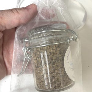 May include: A small glass jar with a silver clasp closure filled with loose brown herbs. The jar is in a white sheer organza bag with a drawstring closure. The label on the jar reads "Gift from the Garden" with a handwritten name and the words "Lavender Mint" in cursive.