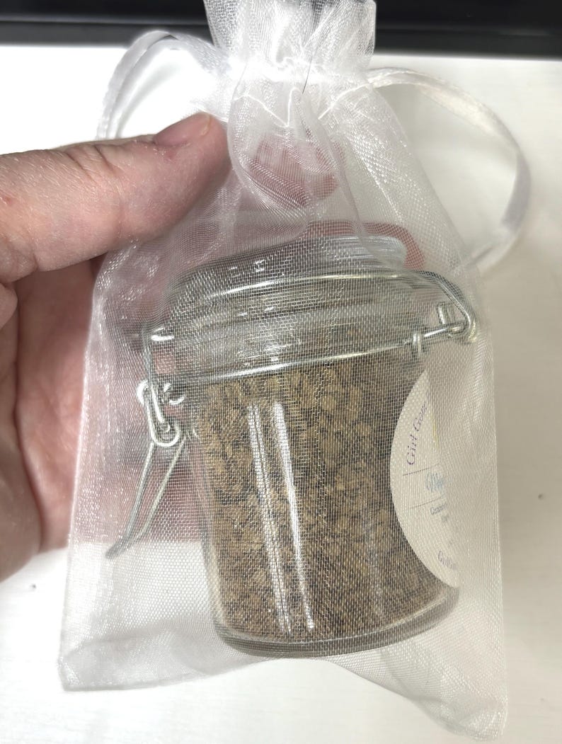 May include: A small glass jar with a silver clasp closure filled with loose brown herbs. The jar is in a white sheer organza bag with a drawstring closure. The label on the jar reads "Gift from the Garden" with a handwritten name and the words "Lavender Mint" in cursive.