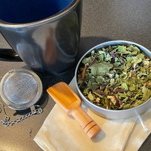 May include: A black ceramic mug with a blue interior, a silver tea infuser with a chain, a wooden scoop, and a metal container filled with dried herbs.