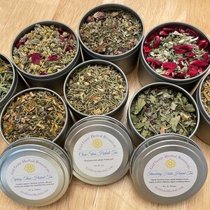 May include: Eight metal tins filled with various dried herbs and flowers. The tins have labels with the text "Gil Gone Herbal Botanicals" and the names of different herbal teas, including "Spring Time Herbal Tea", "Clean Skin Herbal Tea", and "Strawberry Fields Herbal Tea".