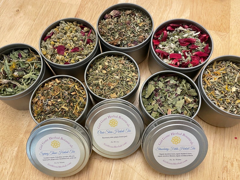 May include: Eight metal tins filled with various dried herbs and flowers. The tins have labels with the text "Gil Gone Herbal Botanicals" and the names of different herbal teas, including "Spring Time Herbal Tea", "Clean Skin Herbal Tea", and "Strawberry Fields Herbal Tea".