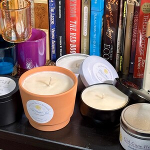 May include: A collection of scented candles in various containers, including a terracotta pot, black tins, and a silver tin. The candles have white wax and visible wicks. The background features books and decorative glass holders.