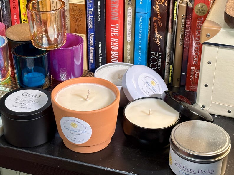 May include: A collection of scented candles in various containers, including a terracotta pot, black tins, and a silver tin. The candles have white wax and visible wicks. The background features books and decorative glass holders.