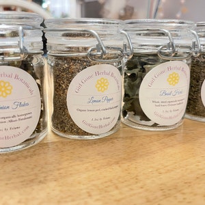 May include: Four glass jars with metal lids containing dried herbs and spices. The labels on the jars read "Green Onion Flakes", "Lemon Pepper", "Basil Herb", and "Thyme Herb". Each label includes the company name "Girl Gone Herbal" and the size of the product, "1.5oz by Volume".