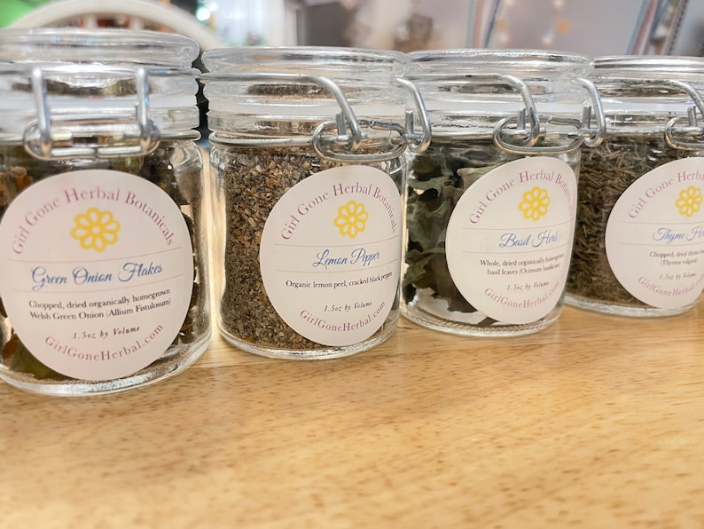 May include: Four glass jars with metal lids containing dried herbs and spices. The labels on the jars read "Green Onion Flakes", "Lemon Pepper", "Basil Herb", and "Thyme Herb". Each label includes the company name "Girl Gone Herbal" and the size of the product, "1.5oz by Volume".