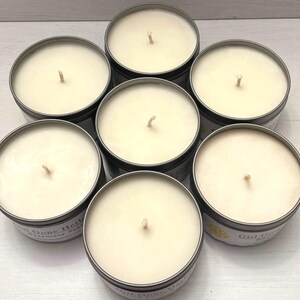 May include: Seven round candles in metal tins arranged in a circular pattern. The candles have a creamy white wax and a single wick. The tins have a dark gray exterior and a label with text. The overall theme is home fragrance.