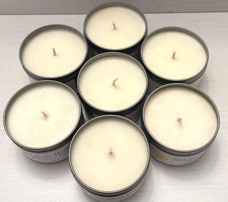 May include: Seven round candles in metal tins arranged in a circular pattern. The candles have a creamy white wax and a single wick. The tins have a dark gray exterior and a label with text. The overall theme is home fragrance.