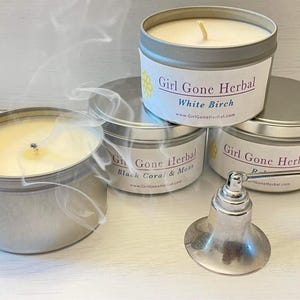 May include: Four silver tin candles with white wax and lit wicks, emitting wisps of smoke. The labels read "Girl Gone Herbal" with scent names "White Birch", "Black Coral & Moss", and "Rose". A silver candle snuffer is in the foreground.