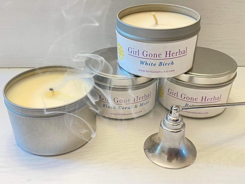 May include: Four silver tin candles with white wax and lit wicks, emitting wisps of smoke. The labels read "Girl Gone Herbal" with scent names "White Birch", "Black Coral & Moss", and "Rose". A silver candle snuffer is in the foreground.