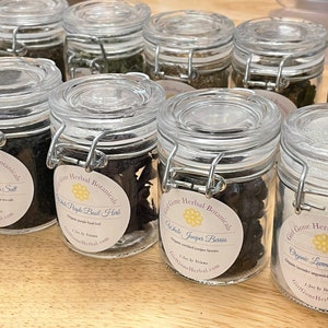 May include: A collection of small glass jars with labels and metal clasps. The jars contain various dried herbs and spices, including black lava sea salt, white pepper basil herbs, white juniper berries, and organic lavender sugar.