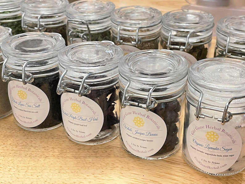 May include: A collection of small glass jars with labels and metal clasps. The jars contain various dried herbs and spices, including black lava sea salt, white pepper basil herbs, white juniper berries, and organic lavender sugar.