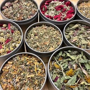 May include: Eight round metal tins filled with various dried herbs and flowers. The tins are arranged in a circle on a wooden surface. The herbs and flowers are a mix of green, brown, red, and yellow.