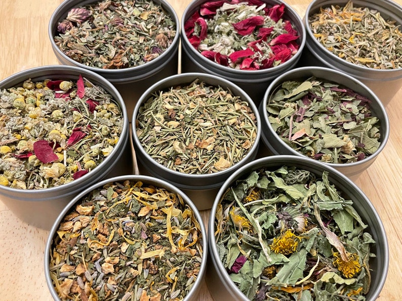 May include: Eight round metal tins filled with various dried herbs and flowers. The tins are arranged in a circle on a wooden surface. The herbs and flowers are a mix of green, brown, red, and yellow.