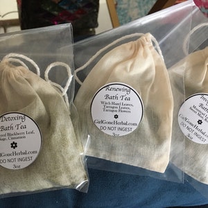 Puede incluir: Tres bolsas de muselina blanca llenas de diferentes tés de baño herbales. Las bolsas están etiquetadas como "Detoxing Bath Tea", "Renewing Bath Tea" e "Invigorating Bath Tea". Cada bolsa tiene una etiqueta con los ingredientes y el sitio web "GirlGoneHerbal.com".