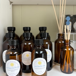 May include: A collection of eight amber glass bottles with black lids and labels. The labels feature a yellow flower and the text "Girl Gone Herbal Botanicals". The bottles are filled with a clear liquid and are arranged on a white surface.
