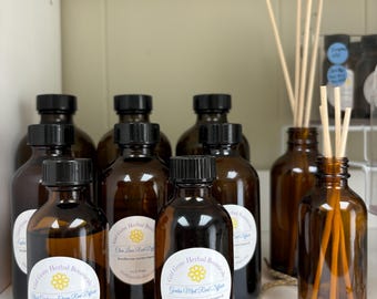 May include: A collection of eight amber glass bottles with black lids and labels. The labels feature a yellow flower and the text "Girl Gone Herbal Botanicals". The bottles are filled with a clear liquid and are arranged on a white surface.