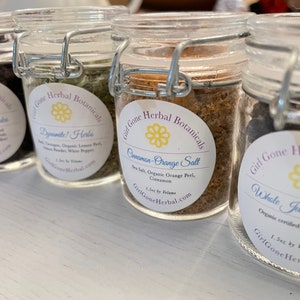 May include: Four glass jars with labels and metal lids containing various herbs and spices. The labels are white with black text and a yellow flower design. The labels read "Girl Gone Herbal Botanicals" and list the contents of each jar: "Red Hibiscus Flowers", "Dynamite! Herbs", "Cinnamon-Orange Salt", and "Whole Juniper Berries".