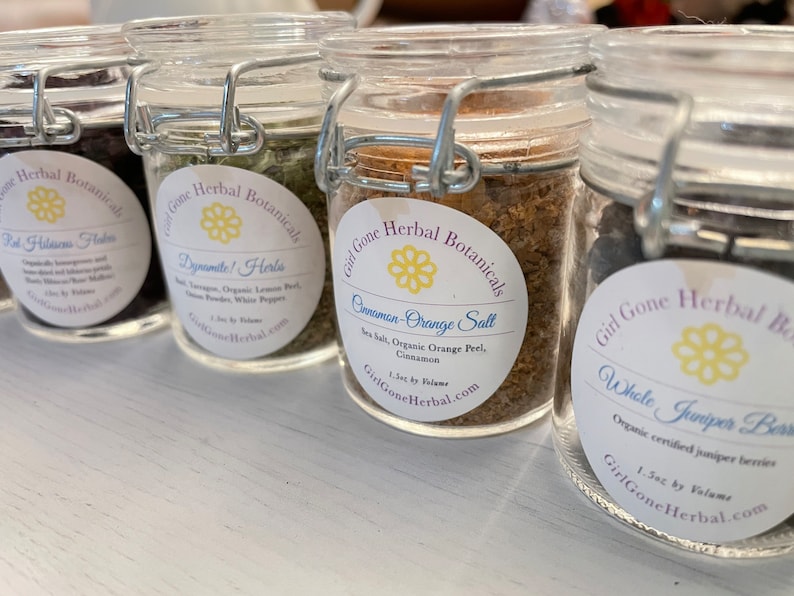 May include: Four glass jars with labels and metal lids containing various herbs and spices. The labels are white with black text and a yellow flower design. The labels read "Girl Gone Herbal Botanicals" and list the contents of each jar: "Red Hibiscus Flowers", "Dynamite! Herbs", "Cinnamon-Orange Salt", and "Whole Juniper Berries".