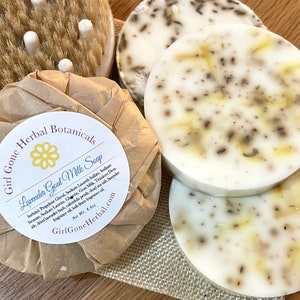 May include: Three round bars of white goat milk soap with dried lavender and other botanicals. The soap is wrapped in brown paper with a yellow flower and the text "Girl Gone Herbal Botanicals Lavender Goat Milk Soap Net Wt. 4 oz. GirlGoneHerbal.com".