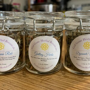 May include: Three glass jars with labels and lids containing dried herbs. The labels read "Sportsman Rub", "Grilling Herbs", and "Dynamite! Herbs". Each jar contains 1.5oz by volume and is labelled "Girl Gone Herbal Botanicals".
