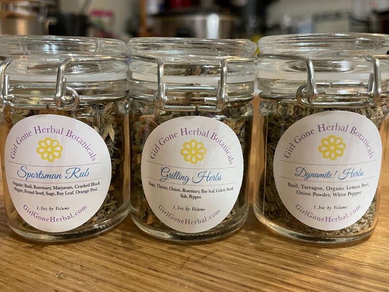May include: Three glass jars with labels and lids containing dried herbs. The labels read "Sportsman Rub", "Grilling Herbs", and "Dynamite! Herbs". Each jar contains 1.5oz by volume and is labelled "Girl Gone Herbal Botanicals".