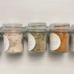 May include: Six small glass jars with metal lids containing various dried herbs and spices. The jars are labelled with the names of the herbs and spices, including "Lemon Orange Salt", "Dried Juniper Berries", and "Vanilla Sugar".