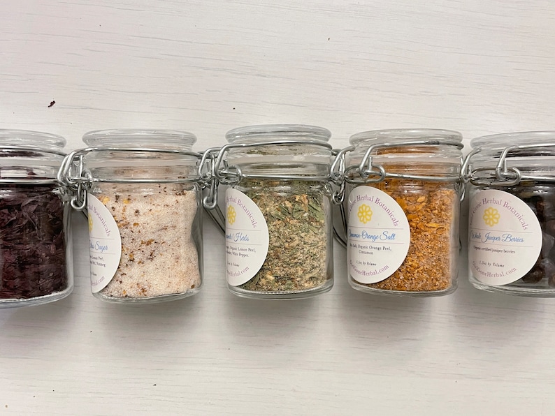 May include: Six small glass jars with metal lids containing various dried herbs and spices. The jars are labelled with the names of the herbs and spices, including "Lemon Orange Salt", "Dried Juniper Berries", and "Vanilla Sugar".