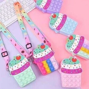 May include: A set of six colorful cupcake-shaped pop-it fidget toys with rainbow, pink, and blue colors. Each toy has a strap for carrying.