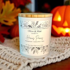 May include: A white candle with a wooden lid, featuring the text "Olive & Mae Candle Co." and "Hocus Pocus." The label includes floral designs and the scent notes: Pomegranate, Lemon Peel, and Cider. The candle is 9 oz (255 g).