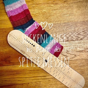 May include: A wooden sock measuring tool with a striped sock on top. The tool has markings for different sock sizes and the text "Sockenl&auml;nge bis Beginn der Spitzen messen" in German.