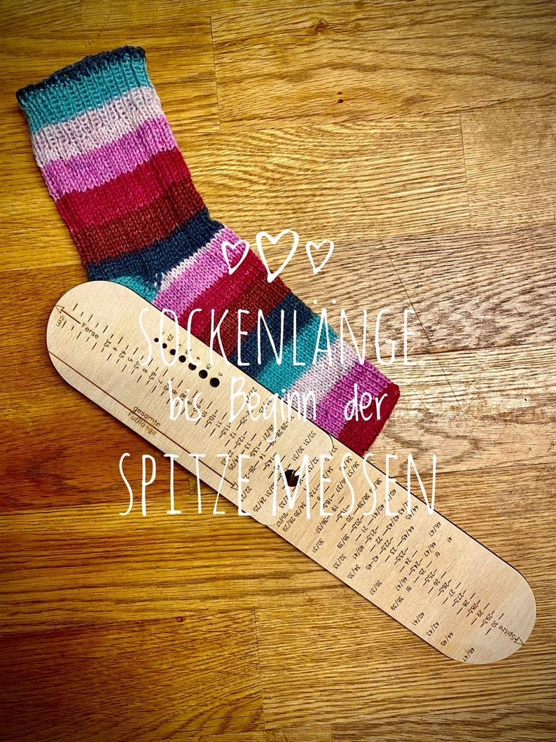 May include: A wooden sock measuring tool with a striped sock on top. The tool has markings for different sock sizes and the text "Sockenl&auml;nge bis Beginn der Spitzen messen" in German.