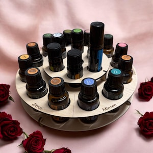 Może przedstawiać: Drewniany stojak ekspozycyjny na olejki eteryczne z dwoma okrągłymi poziomami. Stojak mieści liczne małe, ciemne butelki z kolorowymi etykietami. Większa butelka z kulką znajduje się na środku. Widoczny napis "Zitrus". Czerwone róże w tle.