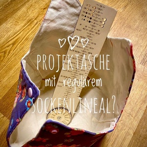 May include: A white fabric project bag with a floral print and pink buttons. Inside the bag is a wooden sock ruler and a wooden sock gauge. The bag has the text "PROJECTASCHE mit regularem SOCKENLINEAL?" printed on it.