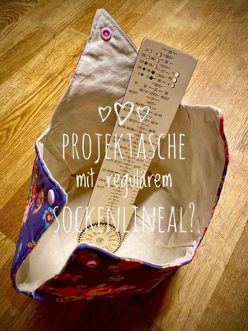 May include: A white fabric project bag with a floral print and pink buttons. Inside the bag is a wooden sock ruler and a wooden sock gauge. The bag has the text "PROJECTASCHE mit regularem SOCKENLINEAL?" printed on it.