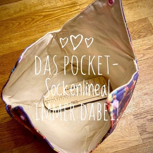 May include: A beige fabric pouch with a floral print lining. The pouch is open and a wooden sock measuring tool is inside. The pouch has a pink button on the top and the text "DAS POCKET-Sockennlineal IMMER DABEI!" is written in white on the fabric.