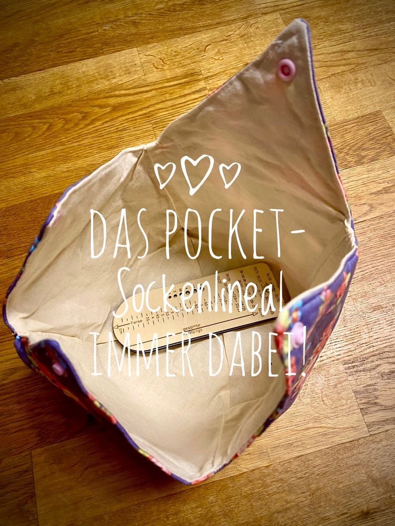 May include: A beige fabric pouch with a floral print lining. The pouch is open and a wooden sock measuring tool is inside. The pouch has a pink button on the top and the text "DAS POCKET-Sockennlineal IMMER DABEI!" is written in white on the fabric.