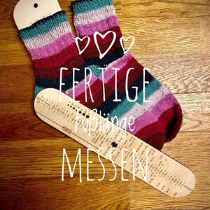 May include: A pair of finished striped socks in shades of pink, red, teal, and black, laid out on a wooden surface with a sock measuring tool. The tool has the text "FERTIGE" and "F&uuml;&szlig;linge MESSEN" printed on it.