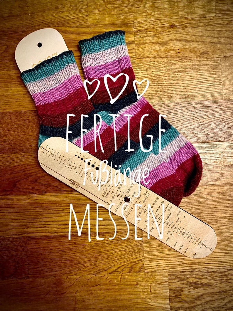 May include: A pair of finished striped socks in shades of pink, red, teal, and black, laid out on a wooden surface with a sock measuring tool. The tool has the text "FERTIGE" and "F&uuml;&szlig;linge MESSEN" printed on it.