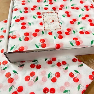 May include: A white cardboard box filled with tissue paper featuring a repeating pattern of red cherries with green stems. A rectangular card with a decorative border and an illustration of a white rabbit is placed on top of the tissue paper.