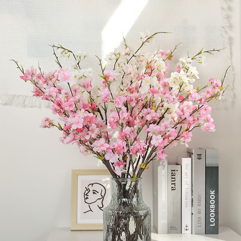 May include: A bouquet of pink and white artificial cherry blossom branches in a clear glass vase with a textured surface. The vase is sitting on a table with a stack of books.
