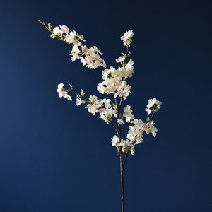 May include: A single white and pink artificial cherry blossom branch with delicate flowers against a dark blue background.