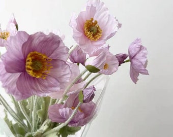 Puede incluir: Un ramo de delicadas amapolas de papel de color rosa pálido con centros amarillos. Las flores están dispuestas en una envoltura de plástico transparente.